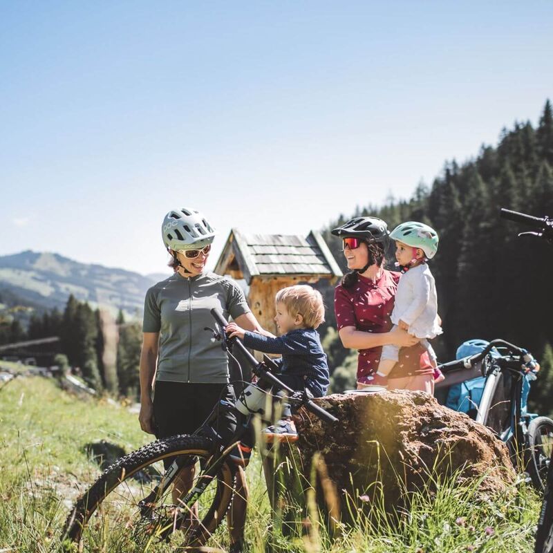 Twee moeders met fietshelmen en hun kinderen nemen een pauze met moderne e-bikes op een alpenweide omringd door berglandschap.