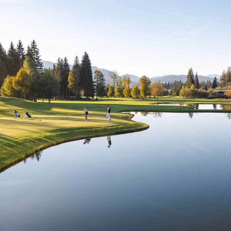Two golfers practice their putting on a tranquil golf course, reflecting autumn colors in the calm water nearby.