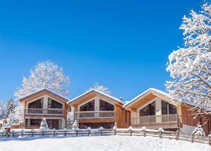 3 Holzchalets mit Balkonen zwischen schneebedeckten Bäumen unter strahlend blauem Himmel