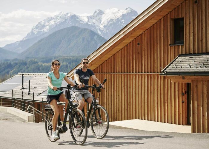 A couple enjoys cycling near a wooden lodge with Schladming's stunning mountain backdrop.