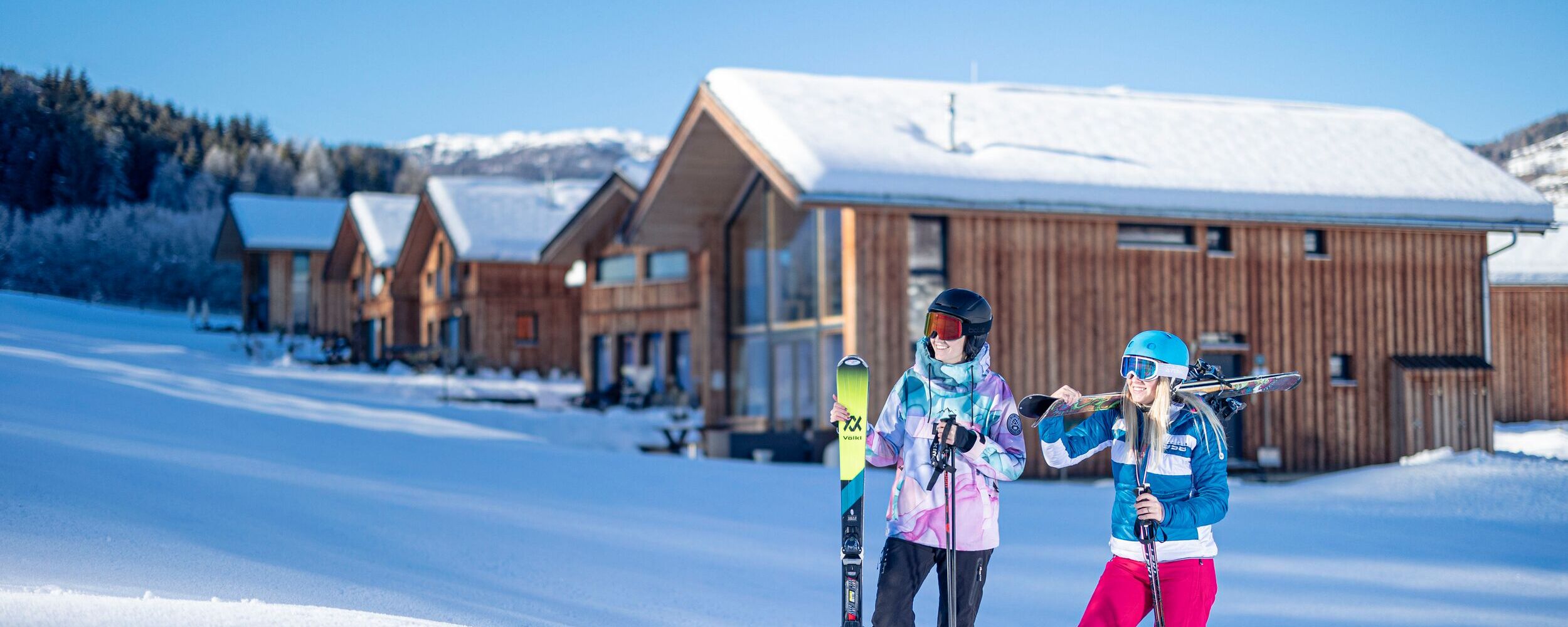 Zwei Skifahrer stehen vor den schneebedeckten Kreischberg Chalets und bereiten sich auf das Skifahren vor.
