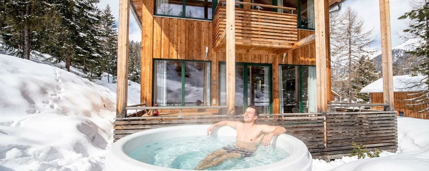 Gast im privaten Hot Tub vor einem Holzchalet in verschneiter Berglandschaft.