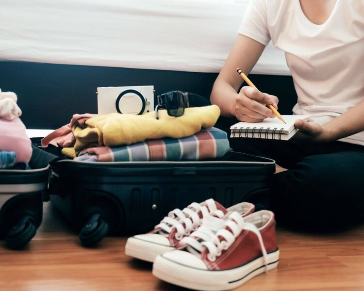 Young woman with a checklist sits on the floor in front of her open, fully packed suitcase.