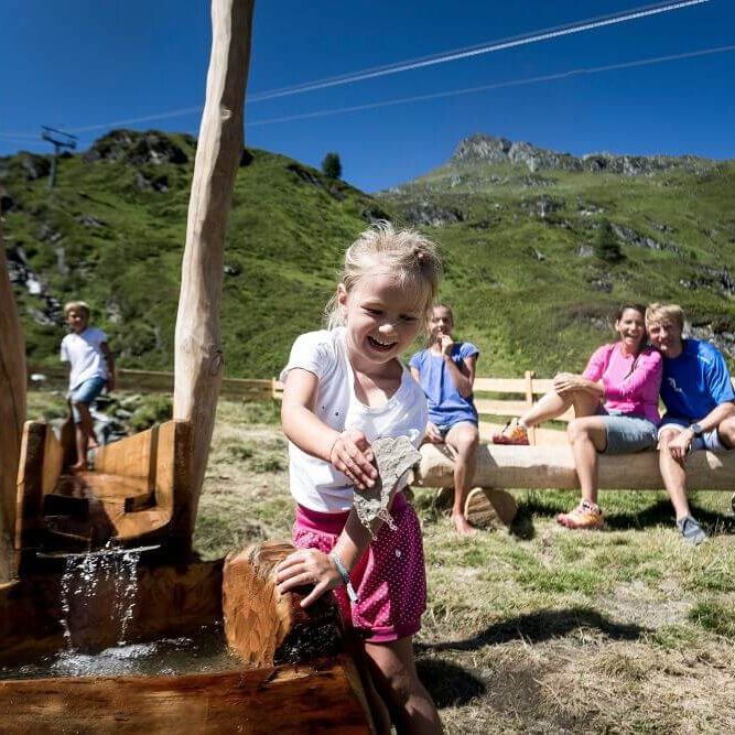 A girl joyfully plays at a water feature while family members relax at a scenic playground. | © Kitzsteinhorn