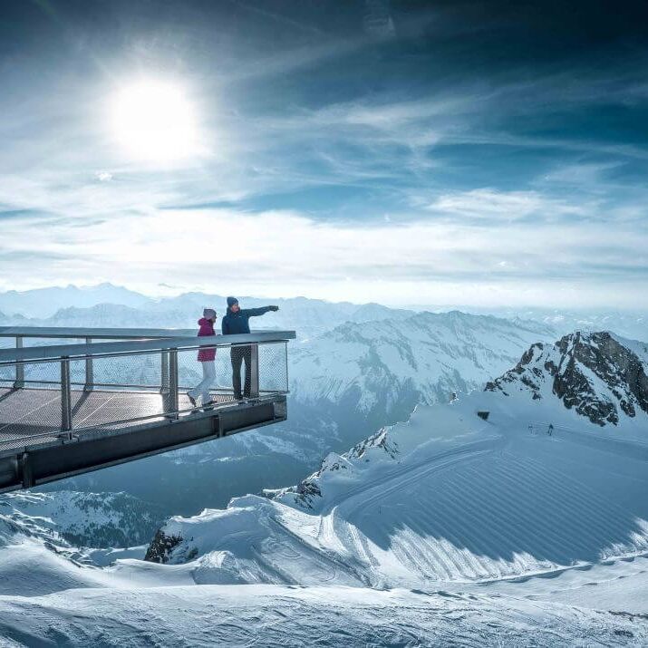 Two visitors enjoy the stunning view from the top of Kitzsteinhorn, surrounded by majestic snow covered mountains.